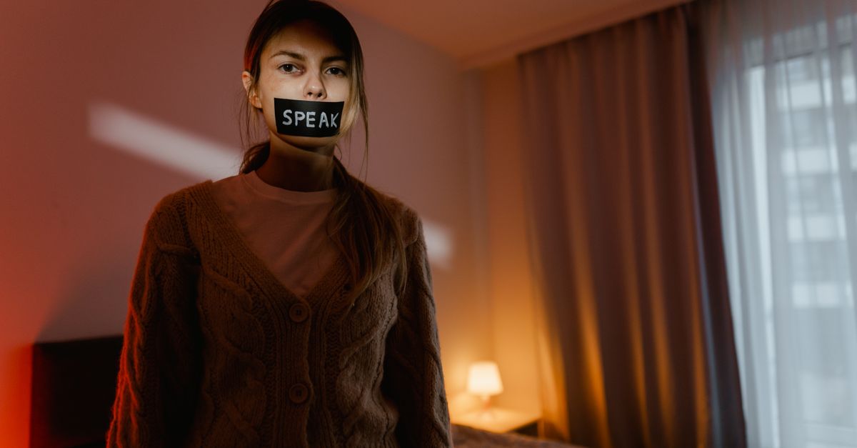 A woman stands in a dimly lit bedroom with tape reading 'Speak' over her mouth, symbolizing silence and oppression.